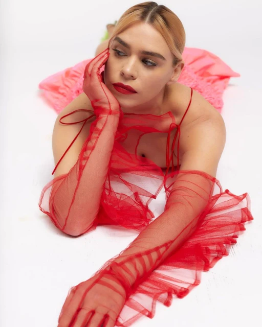 Woman in a red dress and long gloves lies on her side against a white backdrop, gazing sideways with a neutral expression.