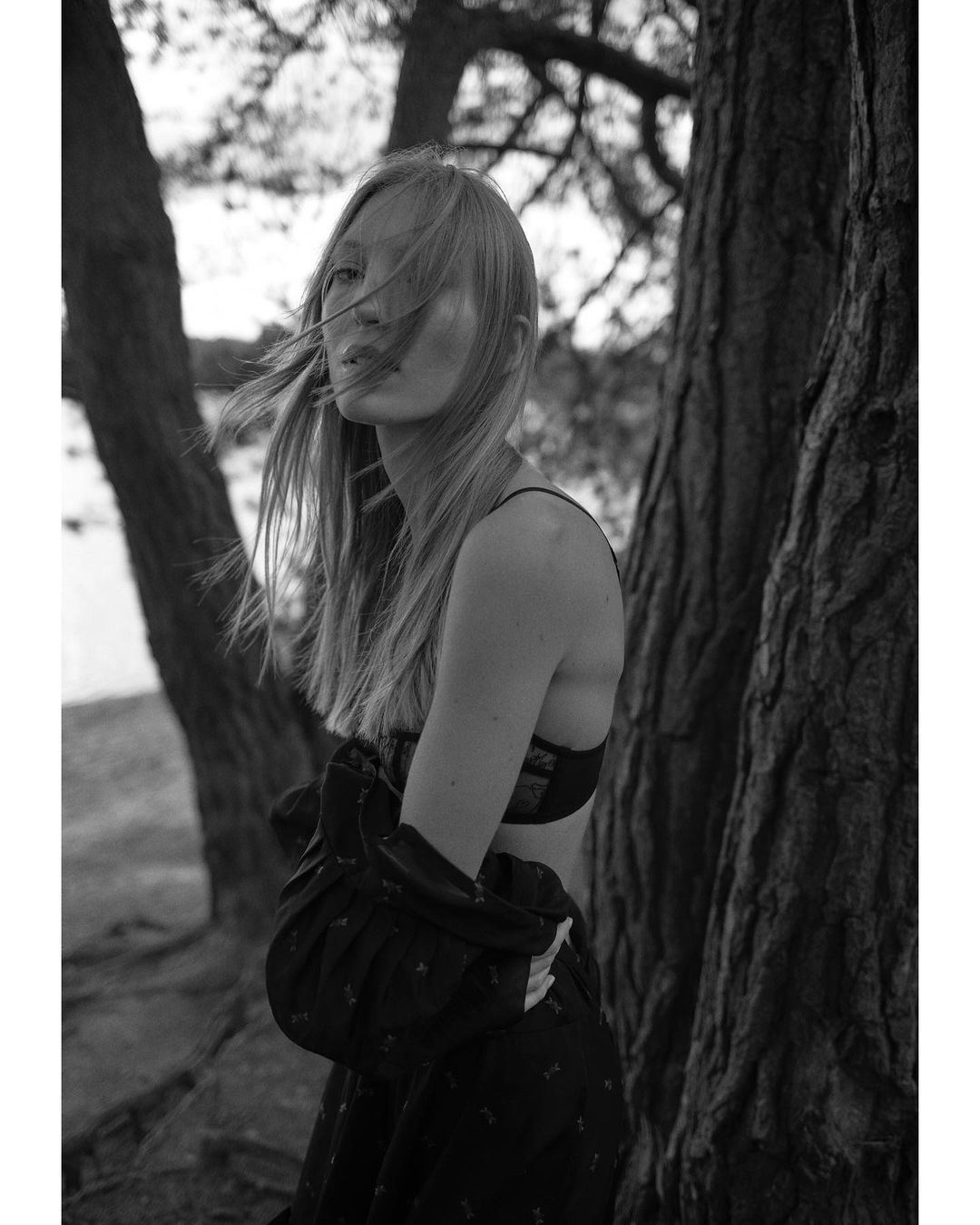 Monochrome portrait of a fair-skinned young woman with long hair, standing among tree trunks in an outdoor environment.