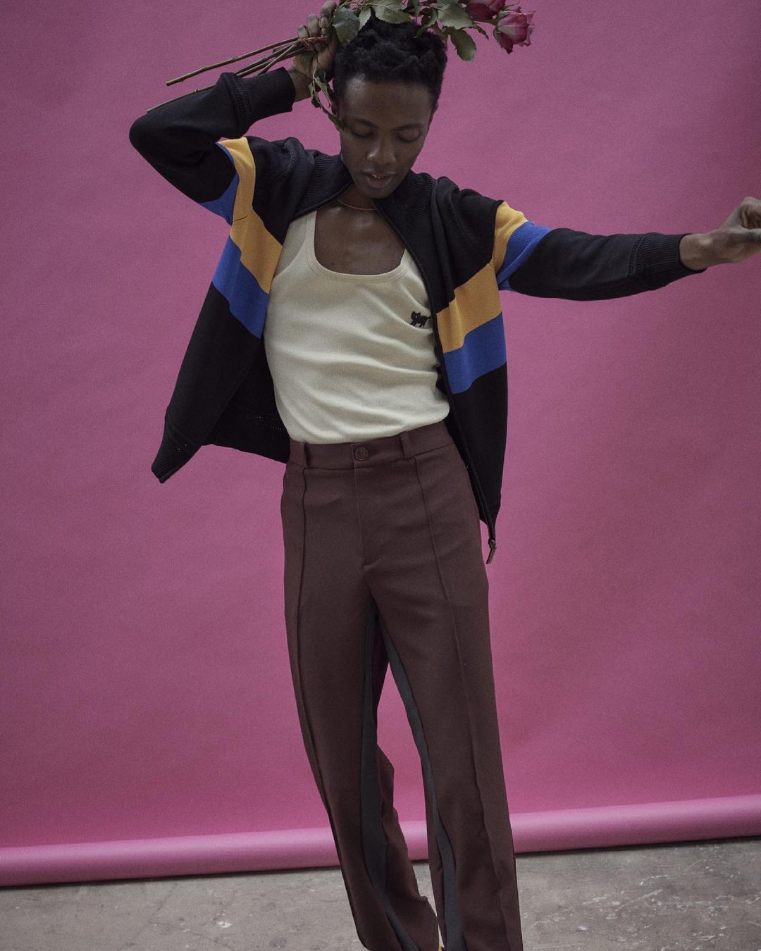 A person poses in a studio wearing a jacket, tank top, and brown pants while holding red roses against a solid pink background.