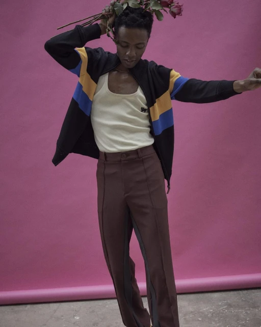 A person poses in a studio wearing a jacket, tank top, and brown pants while holding red roses against a solid pink background.