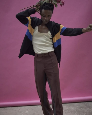 A person poses in a studio wearing a jacket, tank top, and brown pants while holding red roses against a solid pink background.