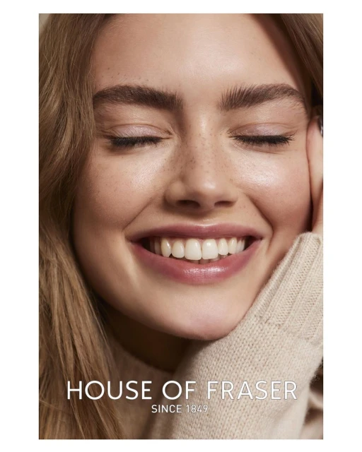 Close-up portrait of a smiling woman with fair skin and light brown hair, wearing a beige sweater, against a plain background.