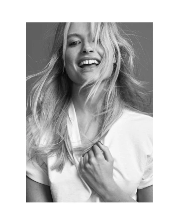 Portrait of a smiling blonde woman with flowing hair wearing a white t-shirt against a plain grey background.