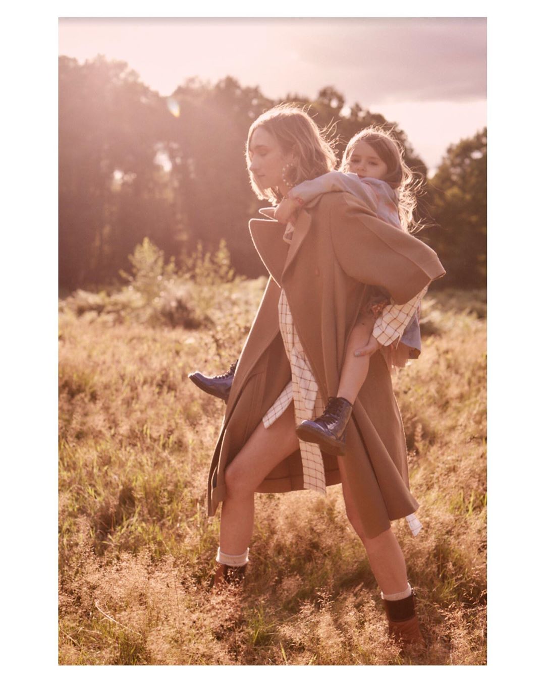 Adult woman carries a young girl on her back outdoors. Both are wearing boots and outerwear in a field of tall grass near trees.