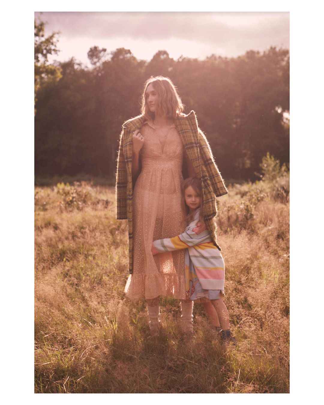 Woman and child in a sunlit field, the woman wearing a plaid jacket and sheer dress, the child in a striped sweater and denim shorts.