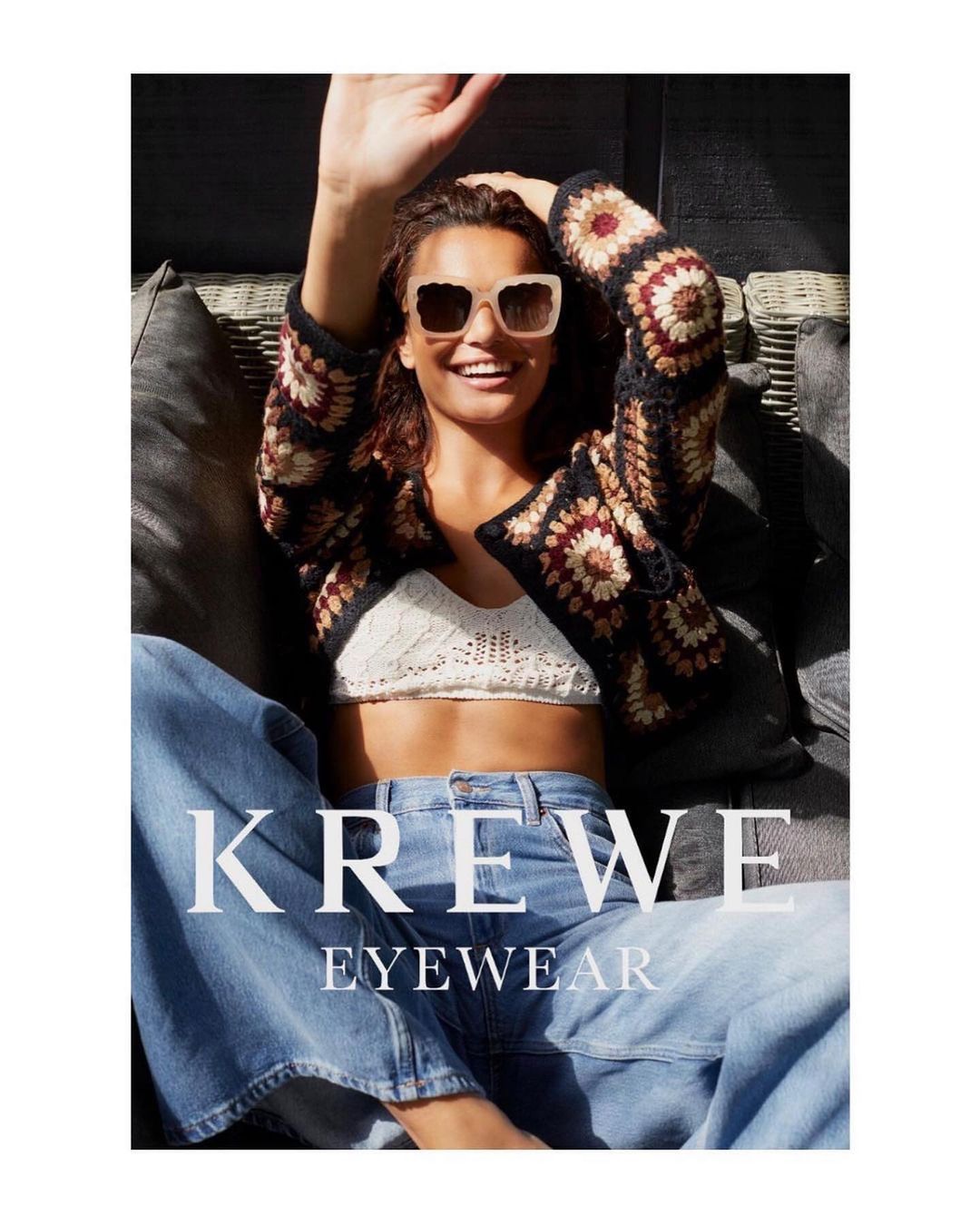 A woman smiles, wearing sunglasses and a crochet jacket, sitting in a relaxed pose with jeans and the KREWE Eyewear logo visible.
