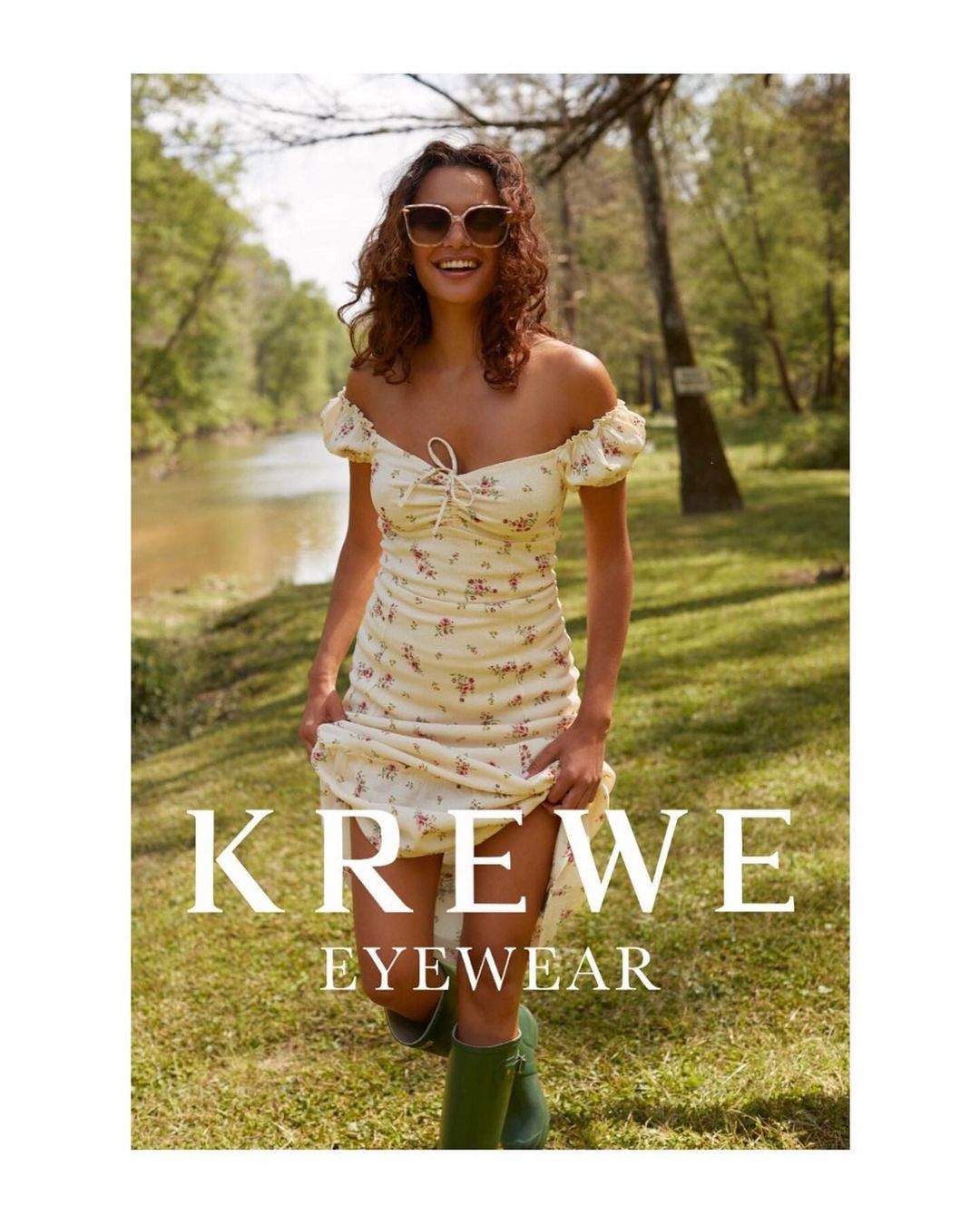 A smiling woman wears a floral dress and sunglasses in a sunny outdoor shot. She is advertising Krewe eyewear while standing in a grassy field.