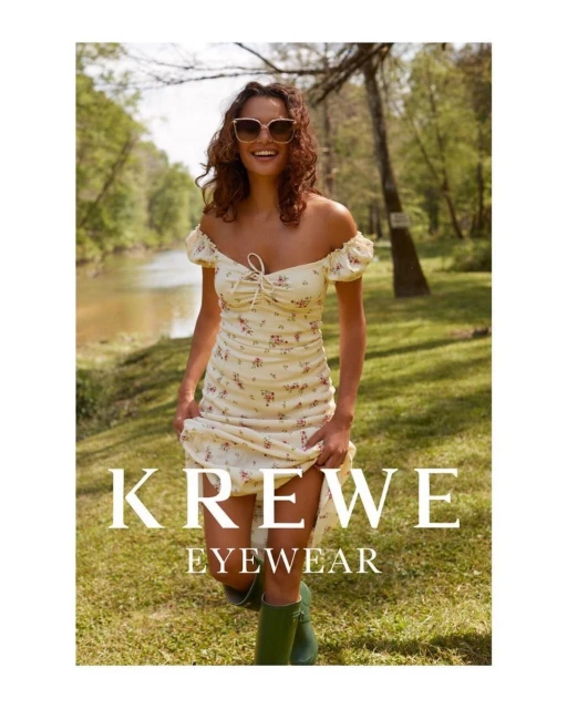 A smiling woman wears a floral dress and sunglasses in a sunny outdoor shot. She is advertising Krewe eyewear while standing in a grassy field.