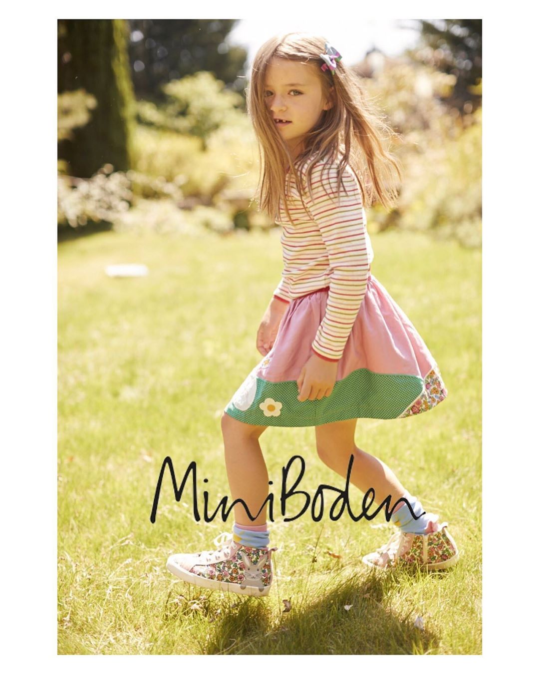 A young girl in a striped shirt and pink skirt walks on a grassy lawn, sneakers visible, Mini Boden logo is visible in the foreground.