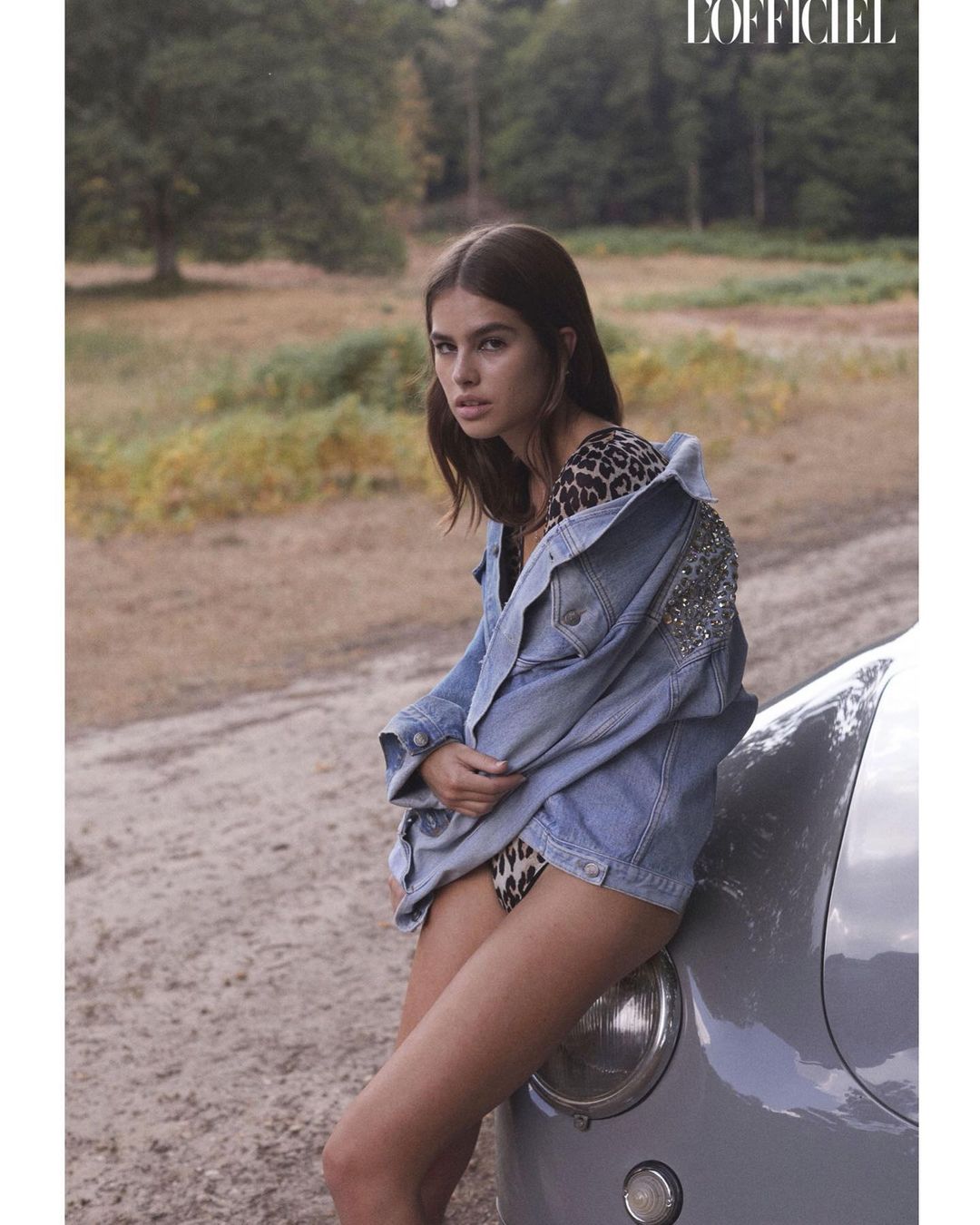 A woman with long brown hair poses with a vintage car in a sun-drenched field, wearing a leopard-print swimsuit and denim jacket.