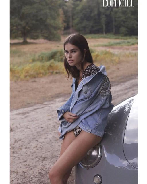 A woman with long brown hair poses with a vintage car in a sun-drenched field, wearing a leopard-print swimsuit and denim jacket.