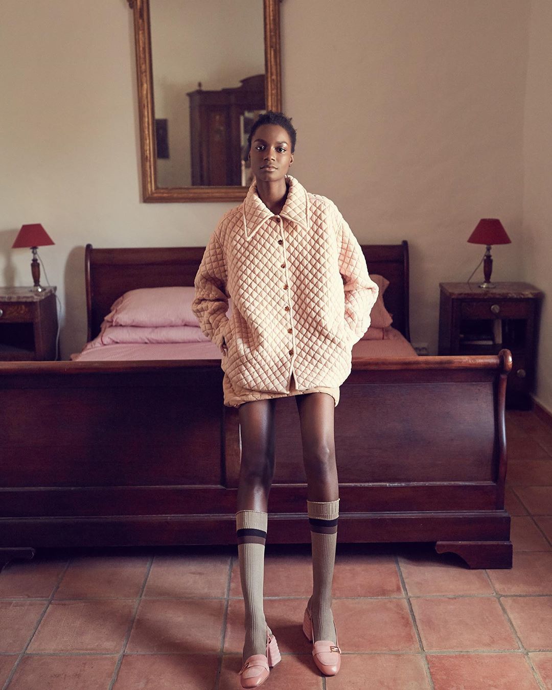 Fashion shot of an African-American woman in an oversized, quilted jacket, socks, and shoes in an indoor bedroom setting.