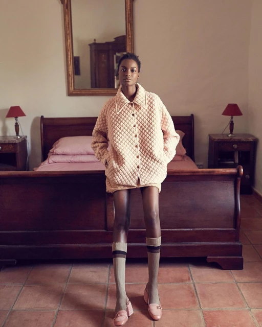 Fashion shot of an African-American woman in an oversized, quilted jacket, socks, and shoes in an indoor bedroom setting.