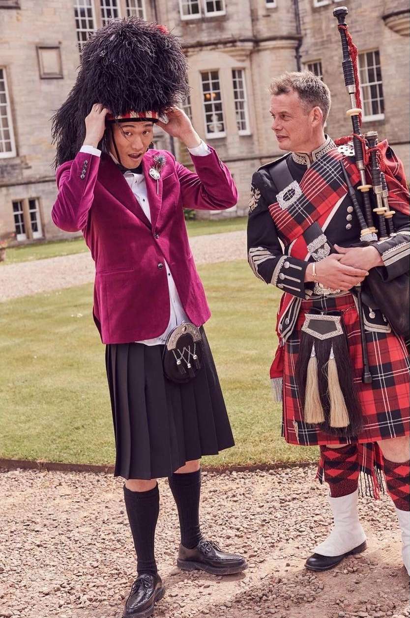 Two men in traditional Scottish attire stand outside a castle; one adjusts a tall black hat, the other holds bagpipes.