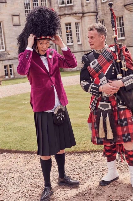 Two men in traditional Scottish attire stand outside a castle; one adjusts a tall black hat, the other holds bagpipes.