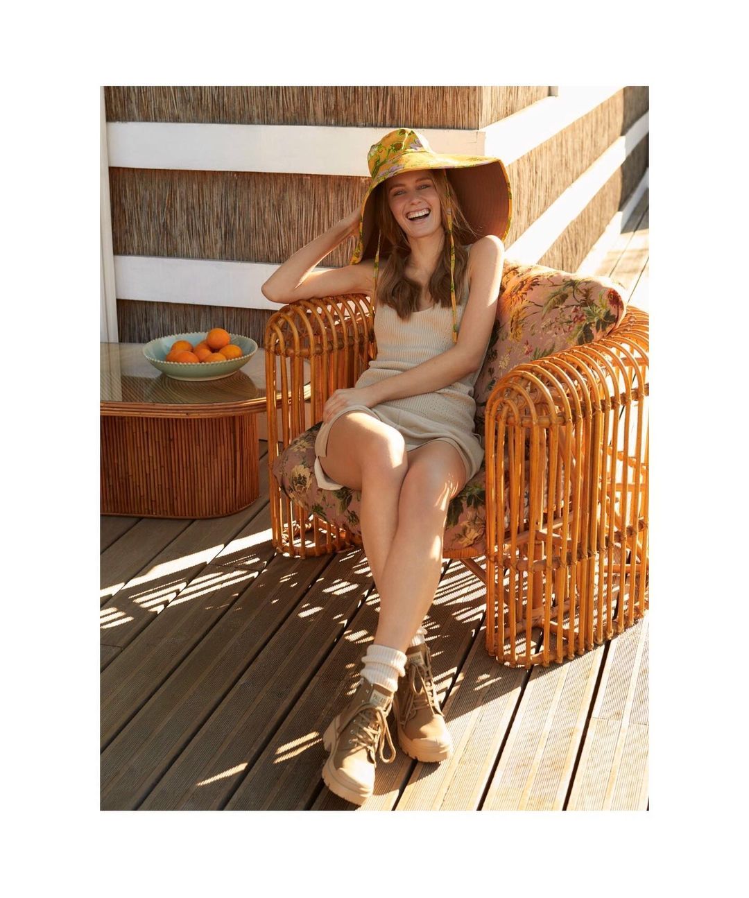 A woman sits in a chair on a wooden porch wearing a large sun hat, a short dress, boots, and socks in the daytime.