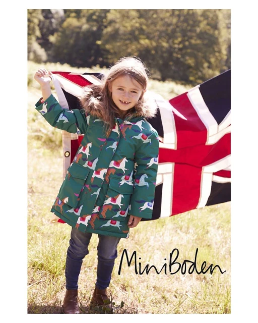 A young girl in a horse-printed coat stands outdoors with a British flag, wearing jeans and brown shoes, smiling at the camera.