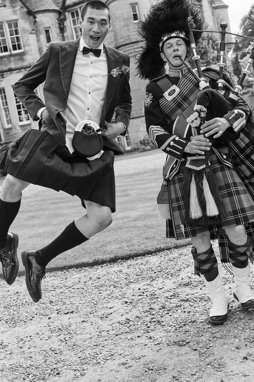 Two men in Scottish kilts, one jumping, another playing bagpipes outdoors near a stone building, in black and white.