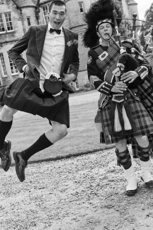 Two men in Scottish kilts, one jumping, another playing bagpipes outdoors near a stone building, in black and white.