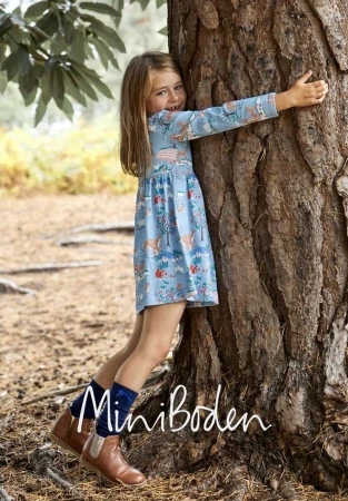 Young girl hugging a large tree trunk wearing a blue patterned dress and brown boots