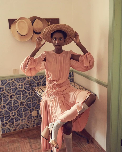 An elegant Black woman in a pink dress and straw hat poses indoors with her leg up, showcasing high-heeled shoes and knee-high socks.
