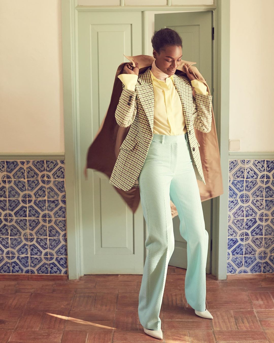 A fashionable Black woman poses indoors wearing a tan blazer, yellow top, light blue pants, and white heels, exuding style and confidence.