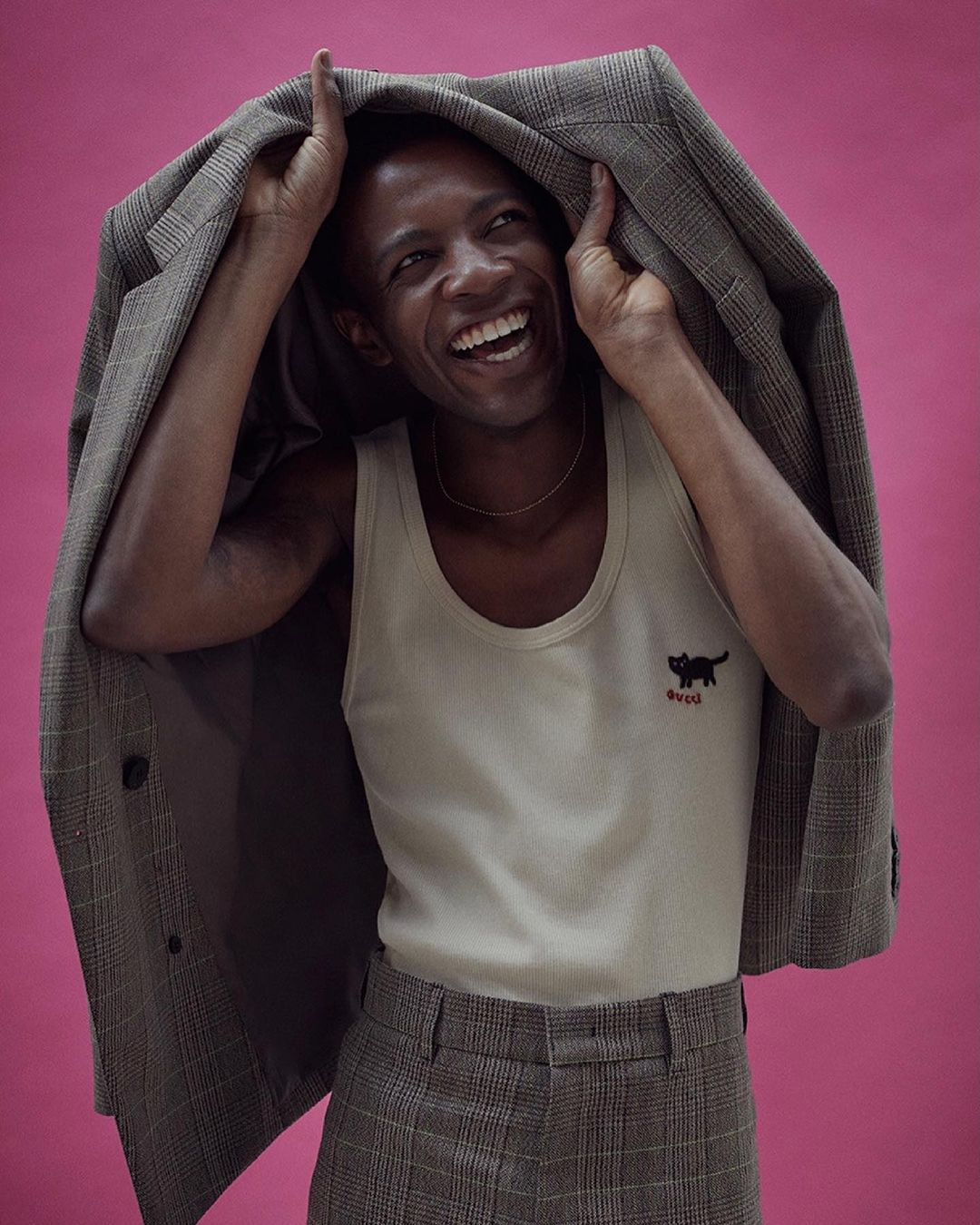 Smiling African male in a tank top and plaid pants holds a plaid blazer over his head against a pink backdrop in a studio shot.