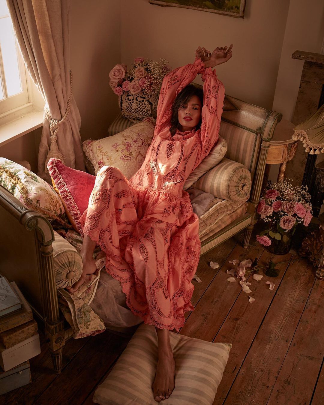 Woman in patterned pink dress reclines on antique chaise lounge amid pillows and rose bouquets in a softly lit, vintage-style indoor setting.