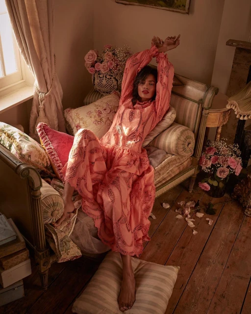 Woman in patterned pink dress reclines on antique chaise lounge amid pillows and rose bouquets in a softly lit, vintage-style indoor setting.