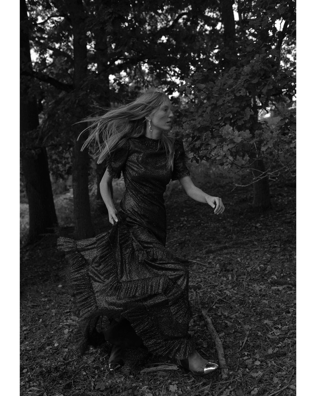 Woman in a sparkly black gown walks in a forest. The image features black and white tones, creating a mood-setting, monochrome scene.