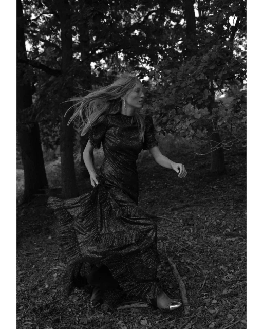 Woman in a sparkly black gown walks in a forest. The image features black and white tones, creating a mood-setting, monochrome scene.