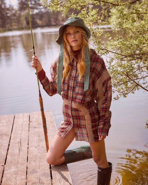 A red-haired woman stands on a wooden dock wearing a plaid shirt and bucket hat while fishing in a lake surrounded by trees.