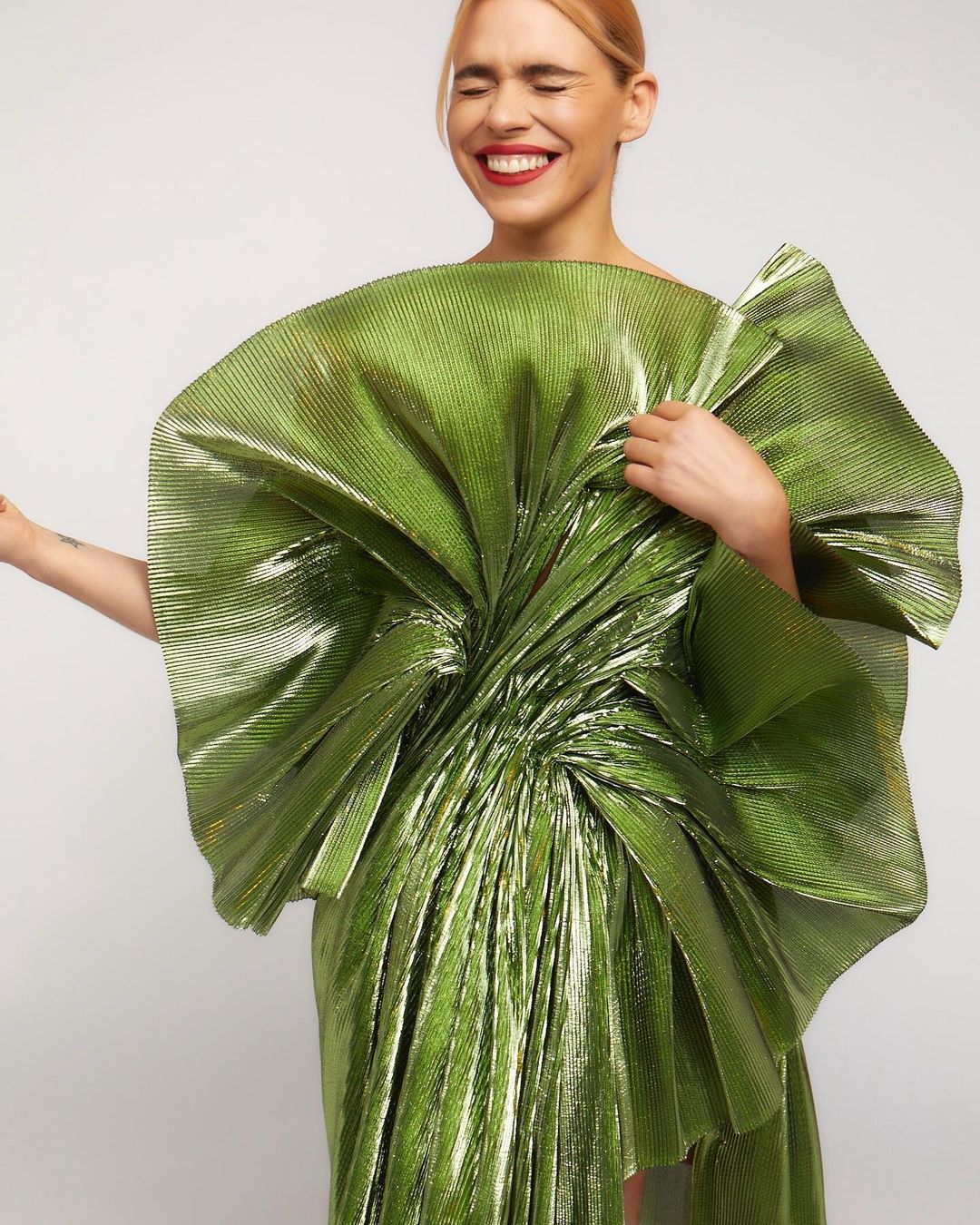 Adult woman in a shiny, large green dress with an oversized bow-like structure, smiling with her eyes closed against a white backdrop.
