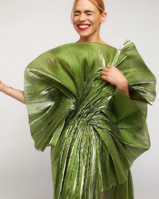 Adult woman in a shiny, large green dress with an oversized bow-like structure, smiling with her eyes closed against a white backdrop.