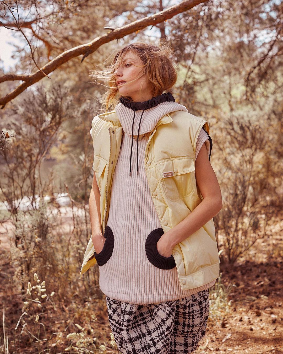 An adult female stands outdoors wearing a puffer vest and sweater, complemented by a plaid skirt and natural sunlight, in a nature setting.