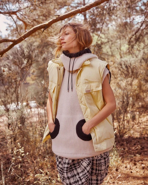 An adult female stands outdoors wearing a puffer vest and sweater, complemented by a plaid skirt and natural sunlight, in a nature setting.
