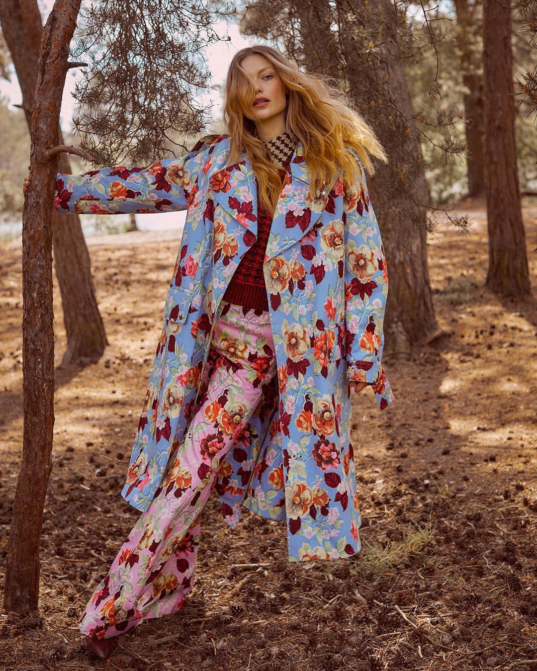 A woman with long blonde hair poses in a forest wearing a floral coat and pants, leaning against a tree on a sunny day.