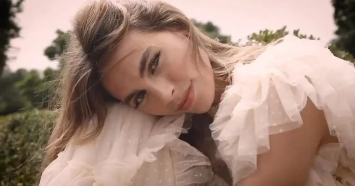 Close-up portrait of a woman with long blonde hair wearing a tiered off-white ruffled dress outdoors.