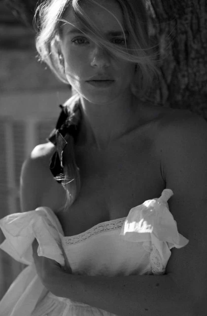 Black and white portrait of a blonde woman with a scarf, wearing a white dress while leaning against a tree outdoors.
