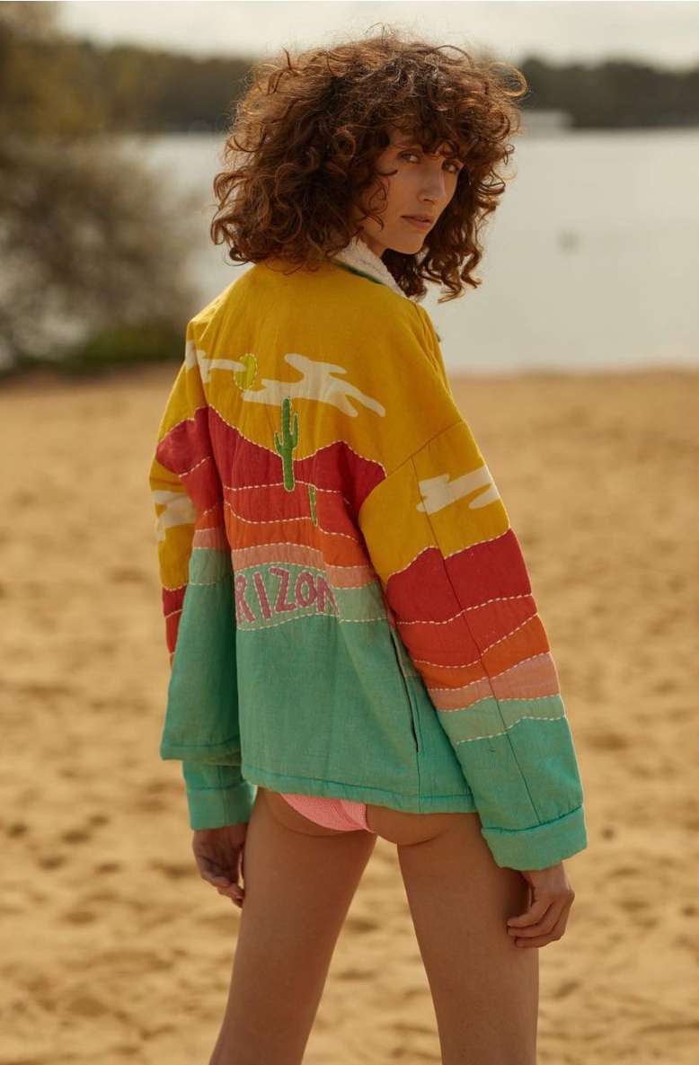 Adult woman wearing an embroidered Arizona-themed jacket on a sandy beach with a lake and trees in the background, looking at the camera.
