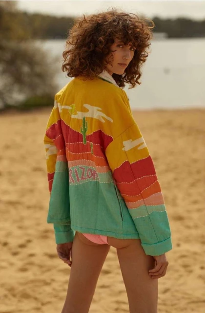 Adult woman wearing an embroidered Arizona-themed jacket on a sandy beach with a lake and trees in the background, looking at the camera.