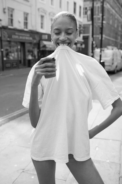 Monochromatic portrait of a smiling adult female on a city street holding a coffee cup and lifting a white t-shirt up to her face.
