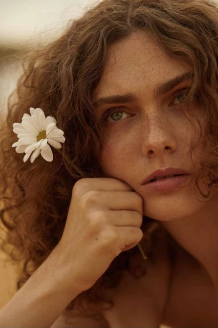 Close-up portrait of an adult female with curly brown hair and green eyes, posing with a white daisy, soft lighting, beauty shot.