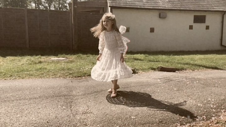 A young girl in a white lace dress with a floral headband is outside on a road next to a white building and grassy areas.