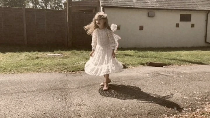 A young girl in a white lace dress with a floral headband is outside on a road next to a white building and grassy areas.
