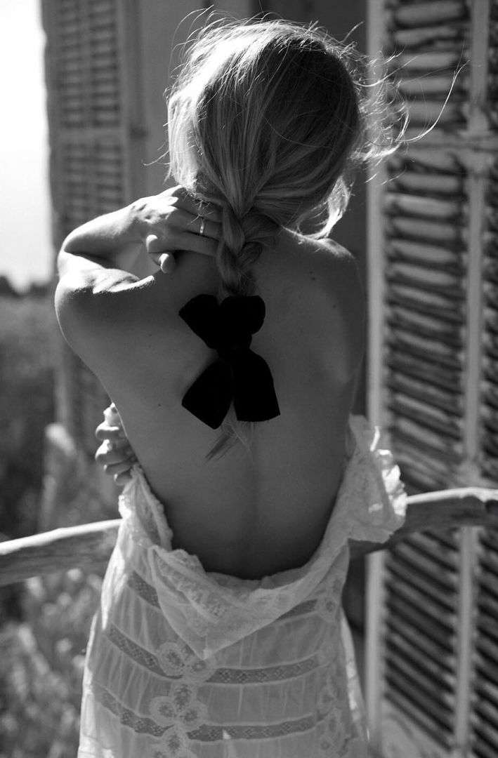 Black and white shot of a woman with blonde hair in a braid, standing on a balcony wearing a lace dress, back to the camera.