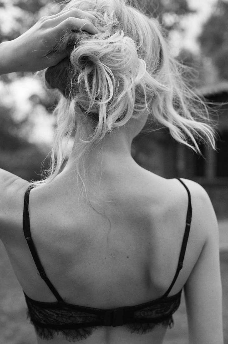 Black-and-white shot of a young adult woman with blonde hair styled in a messy bun, showcasing her bare back and lace lingerie bra.