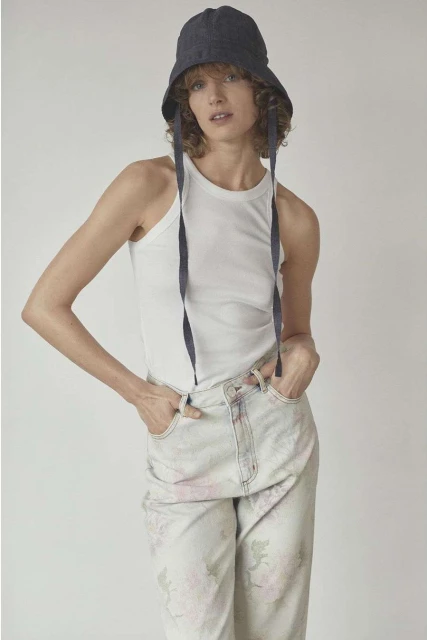 Young woman in a white tank top and patterned jeans poses in a studio setting, wearing a blue bucket hat with dangling ties.