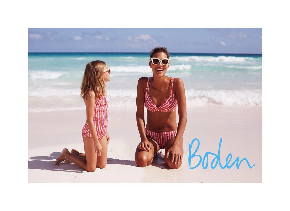 An adult woman and a child, both wearing swimsuits and sunglasses, kneeling on a white sand beach with turquoise ocean in the background.