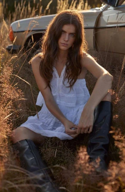 A woman in a white dress and black boots sits in a field next to a vintage car, bathed in golden sunlight during the daytime.
